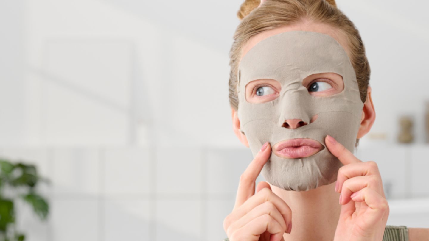 Woman wearing grey sheet face mask pressing cheeks for firming skincare treatment