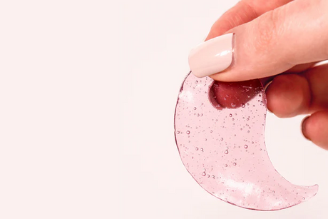 Hand holding pink hydrogel under-eye patch with glossy texture and bubbles on neutral background