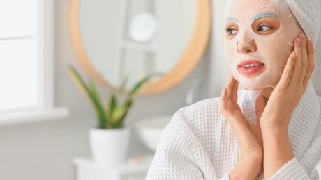 A woman in a white bathrobe with a towel on her hair applies a sheet face mask in a bathroom setting.=