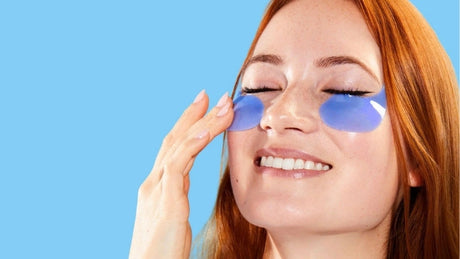 A smiling woman with red hair and closed eyes applying blue under-eye gel patches against a light blue background.
