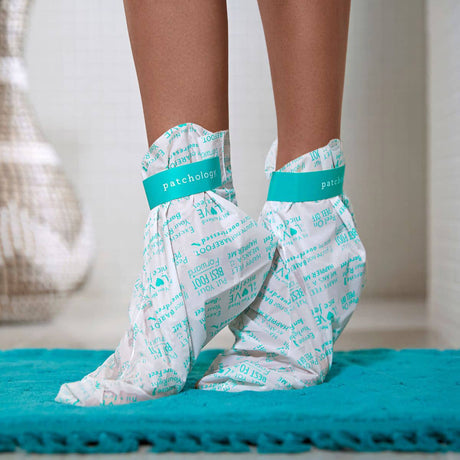 Feet wearing Patchology hydrating foot mask booties on teal bath mat for moisturizing foot treatment
