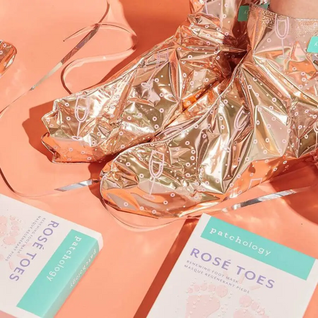 Patchology Rosé Toes foot mask booties in metallic gold with packaging on peach background for hydrating treatment