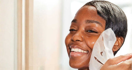 Smiling woman applying a hydrating sheet mask to glowing skin in natural light