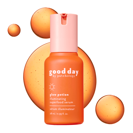Good Day Glow Potion Illuminating Superfood Serum in a 28 mL bottle, featuring a vibrant orange body and a light pink cap, set against a dark green background with abstract orange liquid splashes.