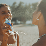 Two women on a beach spraying each other with a blue and white sunscreen bottle, with the text 'that day at the beach' overlaid on the image.
