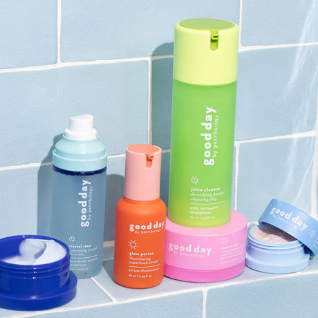 A set of Good Day skincare products arranged on a light blue tiled surface, including a tall green juice cleanser bottle, an orange glow patcher bottle, a blue string cleaner spray, a pink moisturizing cream jar, and a blue cream jar lid.