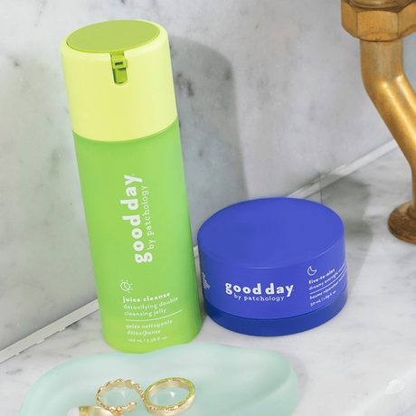 Two Good Day skincare products on a marble bathroom counter: a bright green juice cleanser bottle and a royal blue cream jar, with gold rings on a mint tray nearby.